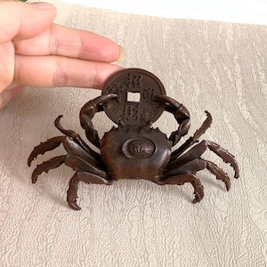 May include: A dark brown metal crab figurine holding a round coin with a square center hole and Chinese characters. The crab has detailed legs and claws, with a small circular emblem on its back. The object is small enough to be held in a hand.