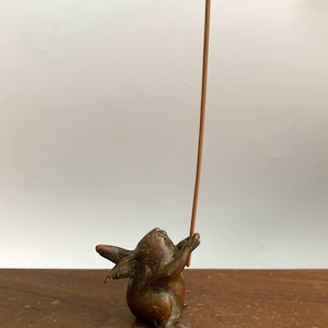 May include: A bronze incense holder shaped like a rabbit, holding a brown incense stick. The rabbit is in a playful pose, looking upwards. The holder is on a wooden surface, with a white background.