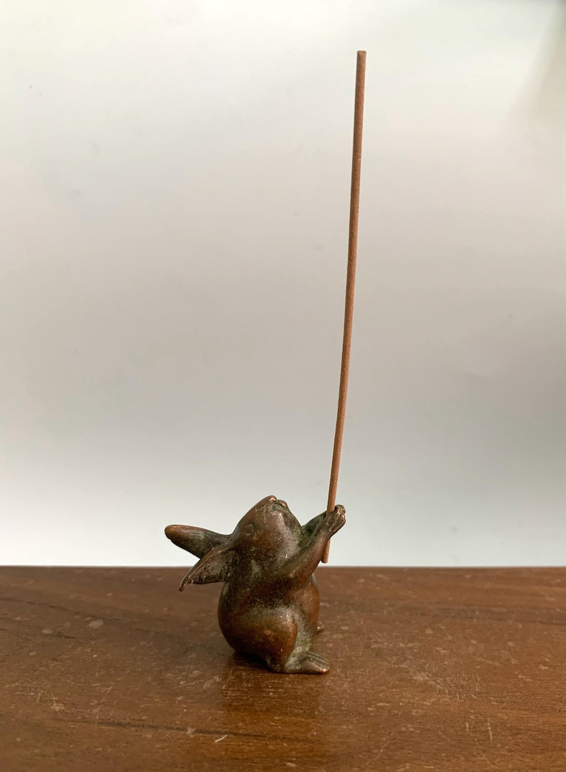 May include: A bronze incense holder shaped like a rabbit, holding a brown incense stick. The rabbit is in a playful pose, looking upwards. The holder is on a wooden surface, with a white background.