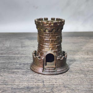 Puede incluir: Una torre de castillo en miniatura de color bronce con un diseño detallado de ladrillos. La torre presenta una parte superior almenada, una pequeña puerta arqueada y una base circular. El diseño recuerda a la arquitectura medieval.
