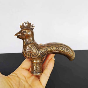 May include: A bronze-colored, bird-shaped decorative handle. The handle features intricate scrollwork and a detailed bird head with a crown. The handle is held in a hand, showcasing its size and design. The handle is likely for a door or drawer.