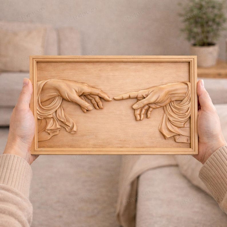 May include: A wooden relief carving of two hands nearly touching, a depiction of Michelangelo's "The Creation of Adam." The artwork is framed in a light brown wooden frame. The hands are sculpted in detail, with flowing drapery.