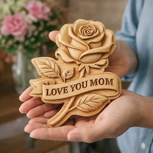 May include: A wooden carving of a rose with the words "LOVE YOU MOM" on a banner. The rose is intricately detailed, with visible petals and leaves. The carving is held in open hands, with a blurred background of pink flowers.