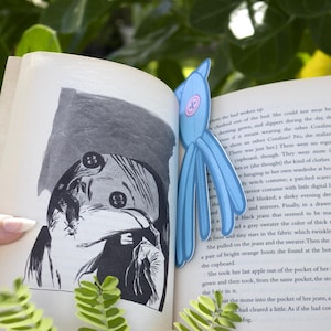 May include: A light blue cartoon-style bookmark shaped like a creature is inserted into a book. The bookmark has a round pink center and long, dangling blue appendages. The book is open to a page with black and white illustrations.