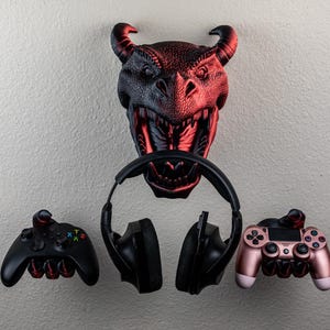 May include: A wall-mounted dragon head with red and black coloring, holding a pair of black headphones. Two game controllers, one black and one rose gold, are mounted below the dragon head. The controllers are designed to hold the headphones.