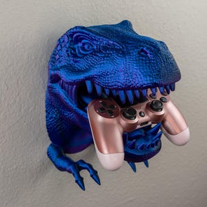 May include: A blue and purple dinosaur head wall mount holding a rose gold and white game controller. The dinosaur has sharp teeth and claws. The wall mount is a unique gaming accessory.