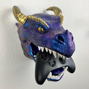 May include: A wall-mounted dragon head sculpture in shades of purple and blue, with gold-colored horns and yellow eyes. The dragon's mouth holds a black video game controller. The sculpture is designed to hold the controller.