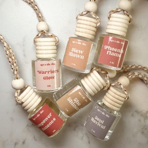 Puede incluir: Una colección de pequeños frascos de vidrio transparente con tapas de madera y cordones beige. Cada frasco tiene una etiqueta con texto como "New Dawn" y "Phoenix Flame". Los frascos están dispuestos sobre una superficie clara.