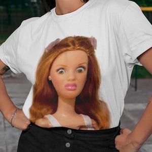 Può includere: T-shirt bianco con stampa di una bambola vintage con espressione sorpresa. La bambola ha i capelli rossi e fiocchi rosa. La maglietta è uno stile casual a maniche corte.