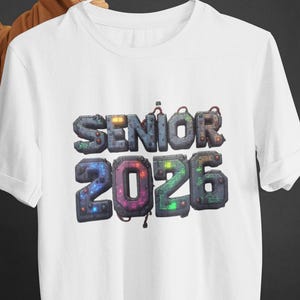 Può includere: T-shirt bianco con la scritta "SENIOR 2026" in un design futuristico 3D. Il testo è in tonalità di grigio, blu, verde e arancione, con una texture simile a un circuito stampato. La maglietta è tenuta da una gruccia bianca.