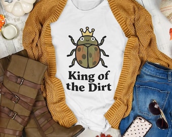 May include: A white t-shirt with a graphic of a beetle wearing a crown and the text King of the Dirt, paired with a mustard yellow open-front cardigan, blue denim jeans, brown knee-high buckled boots, and a white phone case with black dots.