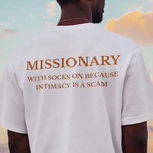 Può includere: T-shirt bianca con la parola "MISSIONARY" stampata in marrone, insieme alla frase "WITH SOCKS ON BECAUSE INTIMACY IS A SCAM". La maglietta è indossata da una persona con i capelli scuri su uno sfondo di cielo.