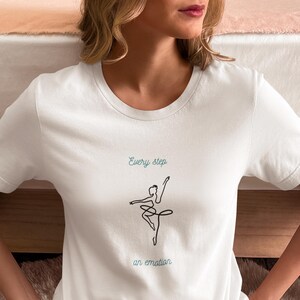 Può includere: T-shirt bianco con un disegno a tratto minimalista di una ballerina e la scritta "Every step an emotion" in turchese. La maglietta è a girocollo ed è realizzata in un materiale morbido.