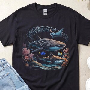 Può includere: T-shirt nero con un'illustrazione dettagliata della vita marina, tra cui uno squalo balena, una manta e altri pesci. Il design incorpora coralli e un banco di pesci, creando una scena sottomarina. La maglietta è abbinata a jeans blu e scarpe da ginnastica bianche.