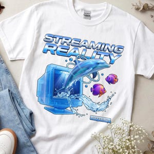 May include: A white short-sleeve t-shirt featuring a graphic print of a blue dolphin leaping out of a vintage computer monitor. The text reads STREAMING REALITY and BUFFERING. Two small purple and orange fish swim nearby amidst blue water splashes.