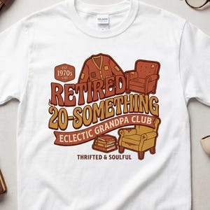 May include: A white short-sleeve t-shirt featuring a graphic print with the text "RETIRED 20-SOMETHING ECLECTIC GRANDPA CLUB" and "THRIFTED & SOULFUL". The design includes illustrations of a cardigan, two armchairs, a stack of books, and a badge.