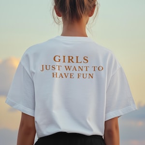Può includere: Una maglietta bianca con la scritta "GIRLS JUST WANT TO HAVE FUN" in marrone. La persona che indossa la maglietta ha i capelli raccolti in uno chignon. Lo sfondo è un gradiente di blu e giallo.