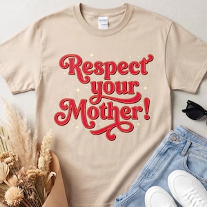 Può includere: Una t-shirt beige a maniche corte con la scritta rossa Respect your Mother! in un carattere stilizzato con piccoli accenti a forma di stella bianca. La maglietta ha un girocollo ed è mostrata insieme a jeans blu, scarpe da ginnastica bianche e fiori secchi.