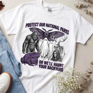Può includere: Una t-shirt bianca a maniche corte con una stampa grafica che riporta il testo PROTECT OUR NATIONAL PARKS OR WE'LL HAUNT YOUR BACKYARD. Il design include illustrazioni di un Bigfoot, una falena, due fantasmi, un cervo scheletrico e un rospo.