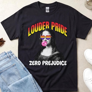 Può includere: Una t-shirt nera a maniche corte con una grafica della Gioconda che indossa occhiali a forma di cuore e una bolla di gomma da masticare rosa. Il testo LOUDER PRIDE appare in un gradiente rosso-giallo sopra e ZERO PREJUDICE è stampato in bianco.