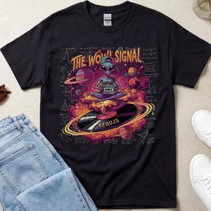 May include: A black t-shirt featuring a graphic of an alien wearing a cap, sitting in a lotus position on a vinyl record, holding a cassette player. The design includes the text THE WOW! SIGNAL and 6EQUJ5, surrounded by planets and math equations.