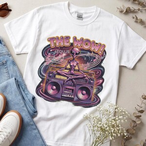 Könnte beinhalten: Ein weißes Kurzarm-T-Shirt mit einer Grafik eines violetten Außerirdischen, der auf einem Ghettoblaster sitzt. Der Text "THE WOW!" steht darüber, daneben "6EQUJ5". Das Design enthält Musiknoten und einen wirbelnden kosmischen Galaxie-Hintergrund.