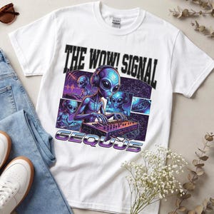 May include: A white short-sleeve t-shirt featuring a graphic print of blue aliens wearing headphones and playing synthesizers. The text reads THE WOW! SIGNAL and 6EQUJ5 in a bold, dark font. The design includes cosmic patterns and purple accents.