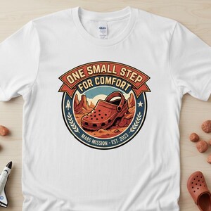 May include: White t-shirt featuring a graphic of a red clog shoe on a Mars-like landscape, with the text "ONE SMALL STEP FOR COMFORT" and "MARS MISSION - EST. 2023". The design includes a rocket and laurel leaves.