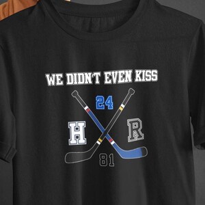 Pode incluir: T-shirt preta com o texto branco "WE DIDN'T EVEN KISS". Abaixo, dois tacos de hóquei cruzados com os números 24 e 81, e as letras H e R. Os tacos de hóquei são azuis e pretos.
