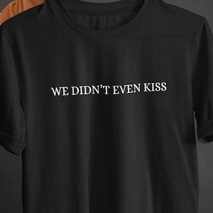 Pode incluir: T-shirt preto com o texto branco "WE DIDN'T EVEN KISS" centrado no peito. A t-shirt de manga curta tem gola redonda e mangas enroladas. A t-shirt é segurada por uma pessoa.