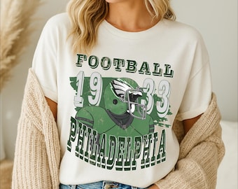 May include: Cream-coloured t-shirt with a green and white graphic. The graphic features a football helmet and the words "FOOTBALL 1933 PHILADELPHIA". The shirt is a casual, vintage-style design.