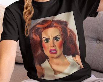 May include: Black t-shirt featuring a doll-like face with red hair, green eyes, and a shocked expression. The doll has bright pink blush and lipstick. The shirt is a crew neck and made of a soft material.