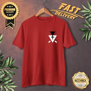 Può includere: T-shirt rosso con una piccola grafica sul petto. La grafica presenta un triangolo nero capovolto sopra un triangolo bianco con una croce e due linee bianche. La maglietta è appesa a una gruccia di legno.