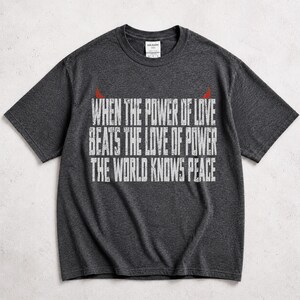 Può includere: Una t-shirt a maniche corte grigio scuro mélange con il testo bianco dall'aspetto usurato "WHEN THE POWER OF LOVE BEATS THE LOVE OF POWER THE WORLD KNOWS PEACE" e due piccole corna da diavolo rosse sopra la scritta sul petto.