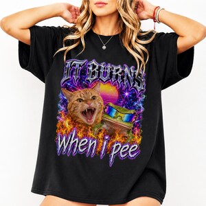 May include: A black t-shirt featuring a graphic print of an orange cat and a green frog wearing sunglasses surrounded by colorful flames and a sunset background. The text IT BURNS WHEN I PEE is printed in a metallic, jagged font above and below the animals.