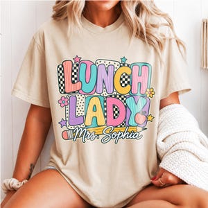 Può includere: Una t-shirt beige con una grafica colorata che riporta la scritta "LUNCH LADY" in grandi lettere stampatello multicolori, seguita dal testo "Mrs. Sophia" sopra l'illustrazione di una matita gialla, con stelle, fiori e una mela.