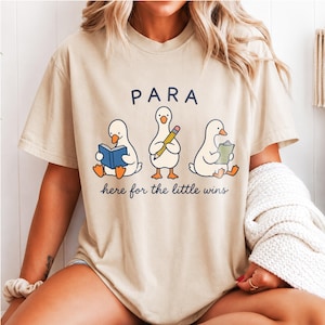 Könnte beinhalten: Ein beiges T-Shirt mit kurzen Ärmeln und einer Grafik von drei weißen Enten. Eine Ente liest ein blaues Buch, eine hält einen gelben Stift und eine hält ein grünes Klemmbrett. Der Text PARA steht darüber und here for the little wins darunter.