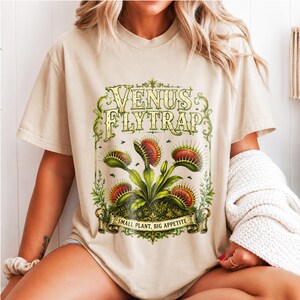 Könnte beinhalten: Ein beiges T-Shirt mit einer grafischen Illustration einer Venusfliegenfalle mit grünen Blättern und rot umrandeten Fallen. Der Text lautet VENUS FLYTRAP oben und SMALL PLANT, BIG APPETITE auf einem Banner unten, umrahmt von verzierten Ranken.