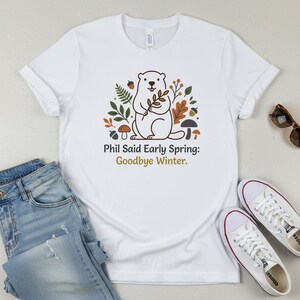 May include: White t-shirt with a cartoon groundhog holding a sprig, surrounded by leaves and mushrooms. Text reads "Phil Said Early Spring: Goodbye Winter." The shirt is styled with blue jeans and white trainers.