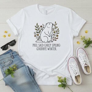 May include: White t-shirt featuring a groundhog illustration with floral accents and the text "PHIL SAID EARLY SPRING: GOODBYE WINTER." The image includes blue jeans, sunglasses, and white trainers, suggesting a casual, everyday style.