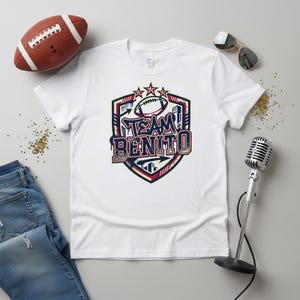 Può includere: Maglietta bianca con una grafica colorata che dice "TEAM BENITO" in un design a scudo. Un pallone da football americano marrone e occhiali da sole sono accanto alla maglietta. Un microfono vintage è anche nell'immagine.
