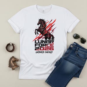 Può includere: T-shirt bianco con grafica di cavallo nero e dettagli rossi, con la scritta "LUNAR FORCE 2026 UNTAMED ENERGY". L'immagine include jeans blu e occhiali da sole. Sono visibili anche una statuetta di cavallo in bronzo e un braccialetto in pelle marrone.