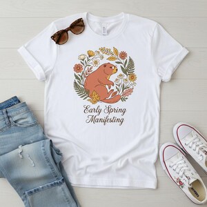 May include: White t-shirt with a brown groundhog design encircled by flowers and the words "Early Spring Manifesting". The shirt is shown with blue jeans, sunglasses, and white trainers.