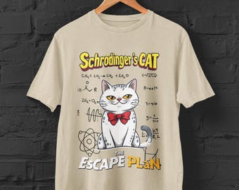 May include: Beige t-shirt featuring a cartoon cat with a red bow tie, labeled "Schrödinger's CAT" and "THE ESCAPE PLAN." The design includes scientific equations and diagrams. The shirt is hanging on a wooden hanger.