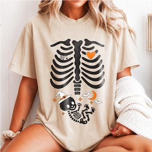 Può includere: Una t-shirt beige a maniche corte con una grafica nera di una gabbia toracica umana con un cuore arancione. Sotto le costole c'è un piccolo scheletro di bambino nero che indossa un cappello da strega bianco, una mezzaluna arancione e pipistrelli.
