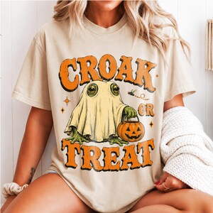 Può includere: Una t-shirt beige con la grafica di una rana verde che indossa un lenzuolo da fantasma bianco e tiene in mano un secchiello a forma di zucca arancione. Il testo CROAK OR TREAT è stampato in grandi lettere arancioni sopra e sotto la rana.