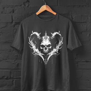 May include: Dark gray t-shirt featuring a white graphic of a skull with horns and intricate, stylized details. The design is centered on the shirt and appears to be a distressed print. The shirt is hanging on a wooden hanger against a dark brick wall.