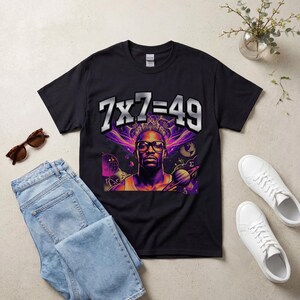 Può includere: T-shirt nero con la scritta "7x7=49" sopra una grafica di una persona con gli occhiali, con un design galattico viola e arancione. L'immagine è presentata con jeans blu e scarpe da ginnastica bianche.