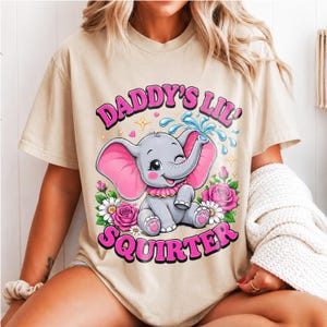 Puede incluir: Una camiseta beige con un gráfico de un bebé elefante gris con orejas rosas, lanzando agua con su trompa. El diseño incluye rosas rosadas, margaritas blancas y el texto DADDY'S LIL' SQUIRTER en letras rosas llamativas con un borde negro.
