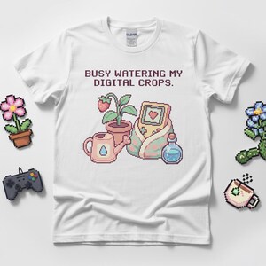 May include: White t-shirt with the text "BUSY WATERING MY DIGITAL CROPS." The shirt features a pixelated graphic of a watering can, a potted plant with a strawberry, a handheld game console, and a potion bottle.